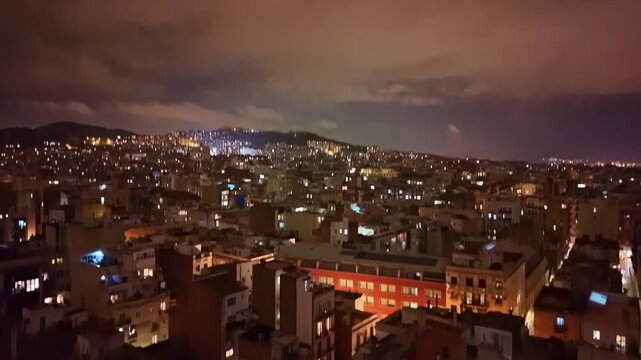 Drone ascent at night over Barcelona, Spain, gradually revealing dense residential rooftops showcasing the vast urban grid expanding toward the Collserola hills beneath a clouded Mediterranean sky.