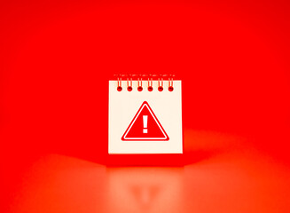 Red warning icon on white spiral desk calendar page over vivid red background. Daily scam alerts,...