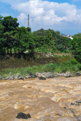 High-angle shot of the Ciliwung River's turbulent brown water rushing through a lush green valley in Bogor, showing the raw power of nature and natural debris after heavy rain in the upstream