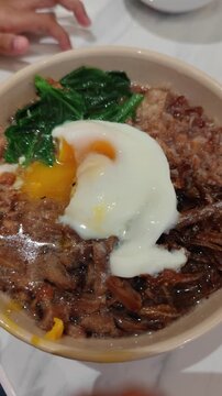 Portrait-oriented video captures a comforting bowl of rice porridge, also known as congee, featuring a soft-boiled egg with a runny yolk, leafy greens, minced meat, and mushrooms in a savory broth. Th
