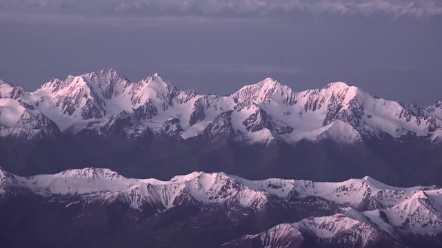Aerial view of snowy Alps peaks in slanting sunlight, winter mountain landscape. Airplane window shows ice covered summits across Europe, glowing at sunrise above clouds.