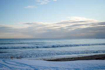 A serene scene of a snowy Pacific coastline with black volcanic sand covered in ice, featuring gentle waves lapping at the shore.