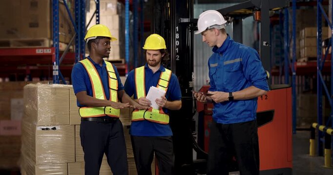 Supervisor instructing two male warehouse workers wearing hardhat and vest while analyzing logistics report in front of forklift beside wrapped shipment boxes inside modern stock facility