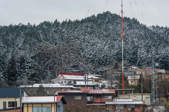 Traditional Japanese houses nestled on a snowy hillside with a tall red and white communication antenna and evergreen forest.