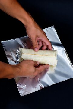 Hands wrapping fresh burrito with foil for delicious homemade meal preparation on dark background for Burrito Day celebration