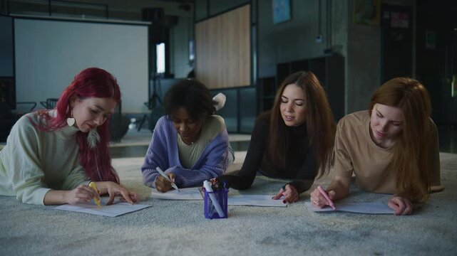 Four women engage in a psychology session, writing and reflecting on personal topics while lying on the floor in a modern indoor location.