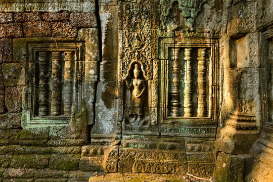 Intricate bas-relief of an apsara dancer and detailed stone window columns are illuminated by warm sunlight on the weathered wall of Ta Nei temple in Angkor, Cambodia. The ancient Khmer masonry