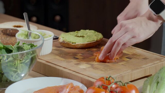 Woman slicing salmon fillet with knife on wooden cutting board with avocado spread on bread and white ramekin in background. Concept of healthy sandwich preparation