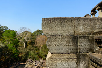 A detailed view of an ornately carved sandstone cornice at Baphuon temple in Angkor, Cambodia. Crisp sunlight highlights the fine Khmer motifs and geometric patterns, with lush forest and clear blue © Florent