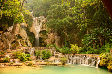 Warm sunlight glows over the tiered limestone cascades and turquoise pools of Kuang Si Waterfalls in northern Laos. Dense tropical forest surrounds the flowing water, highlighting lush greenery and © Florent
