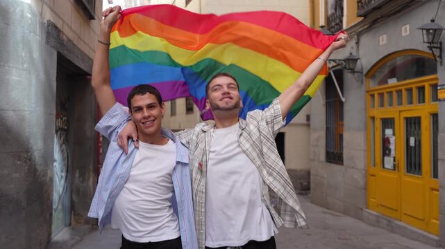 Cheerful young gay couple proudly holding a large rainbow flag while walking and smiling in a narrow city street, celebrating love and pride