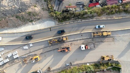 Aerial view of road construction site with heavy machinery, trucks and workers preparing materials beside a busy highway, symbolizing urban infrastructure growth and progress © DawDunia