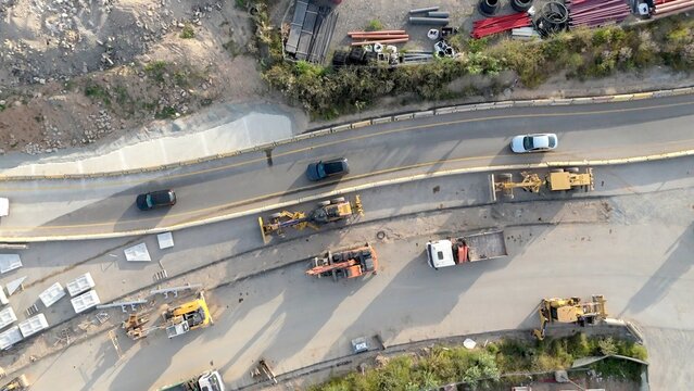 Aerial view of road construction site with heavy machinery, trucks and workers preparing materials beside a busy highway, symbolizing urban infrastructure growth and progress