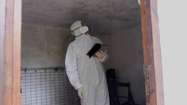 Person in protective suit with respirator mask documenting hazardous conditions, inspecting interior ceiling for mold damage and environmental remediation issues with a tablet