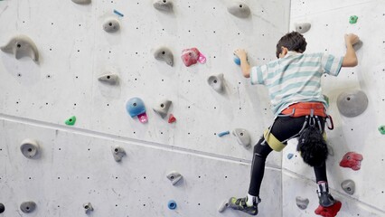 Young boy climbing an indoor artificial wall in harness and climbing shoes, building strength, coordination and focus during a recreational training session in a bright gym environment © DawDunia