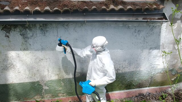 Worker in protective suit and respirator spraying biocidal disinfectant on a grimy exterior wall, demonstrating professional sanitization and decontamination for health and safety