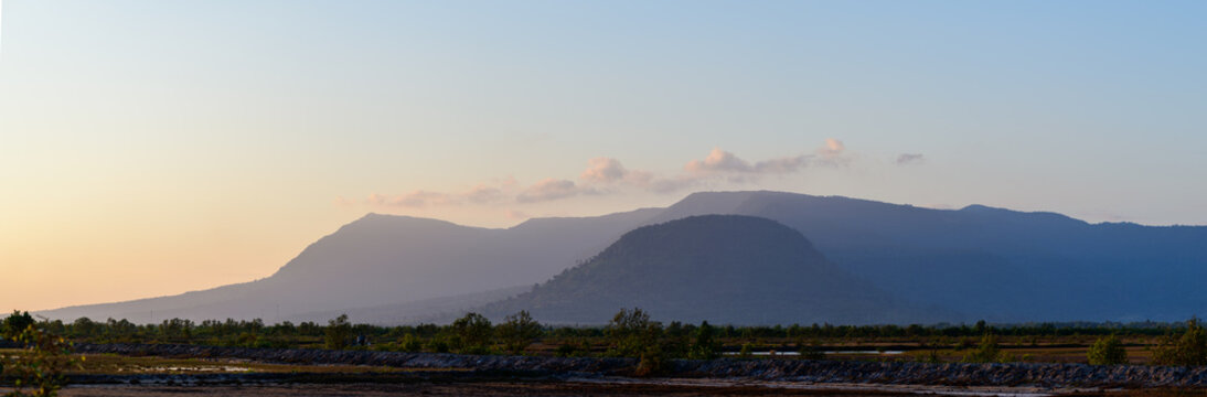 Gentle sunset hues illuminate the layered silhouette of the Bokor Mountains near Kampot, Cambodia. The rural foreground features low vegetation and salt marshes, with subtle clouds drifting above the