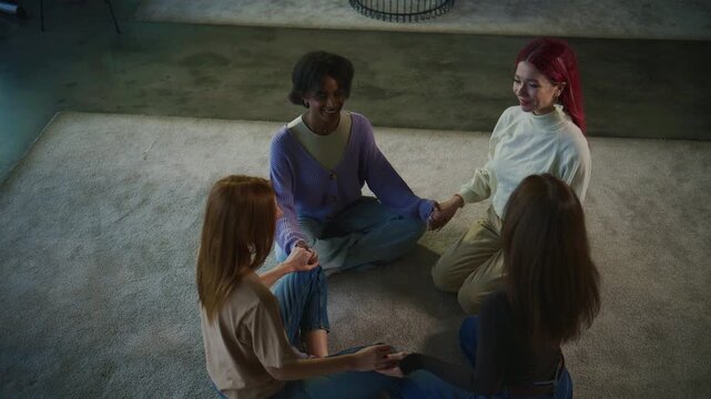 Four women sit on a soft carpet in a room, holding hands and engaging in a discussion during a psychology session aimed at personal development