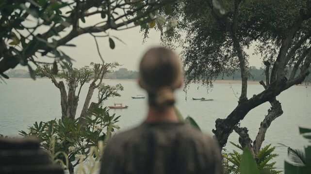 Back view of blonde woman, slightly out of focus, gazes in between trees over the ocean where traditional Balinese fishing boats float on the water.