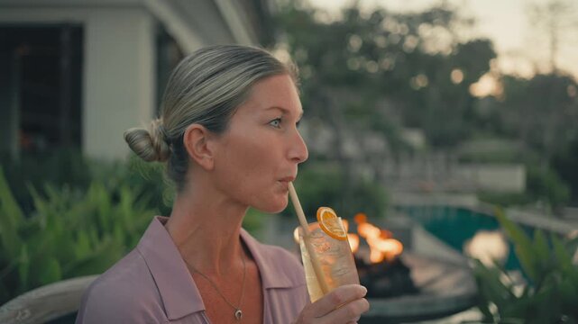 Close up of happy, elegant young blonde woman sipping cocktail at a luxury resort beach club, with a glowing fire pit in the background creating a warm sunset atmosphere