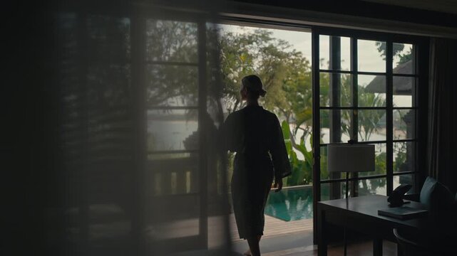 Woman steps outside a luxury pool villa with ocean view, pausing at pool to take in the stunning scenery, framed by a sheer curtain in a cinematic shot