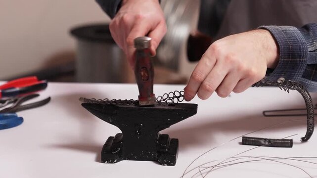 Close up of craftsman shaping metal wire on small anvil using hammer. Traditional Slovak wire craft technique, handmade folk art and heritage craftsmanship in workshop.