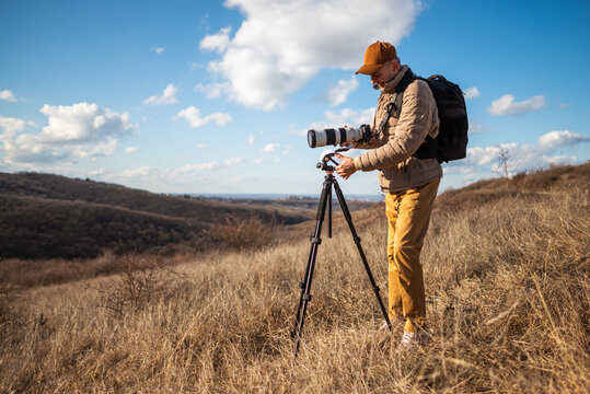Wildlife senior photographer using tripod and setting a large telephoto lens on an open countryside landscape on sunny day.
