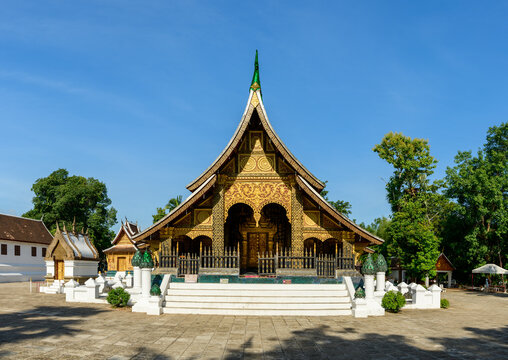 Striking view of an ornate golden Buddhist temple with intricate carvings and sweeping rooflines in Luang Prabang, Laos. The structure stands under clear blue sky, framed by lush green trees and white