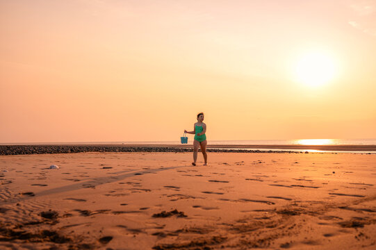 Girl walking with sand bucket along beach at sunset.