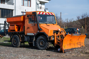 a heavy-duty, bright orange snowplow truck equipped with a large front blade and a rear salt spreader, parked on a gravel lot in front of a white residential building.