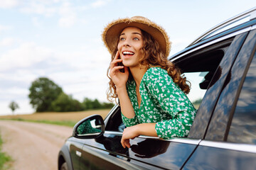Naklejka premium Cheerful woman leans out window, enjoying rural landscape on sunny day. Young traveler in bright dress and hat feels free, catching wind in hair from car window. Concept: transportation, adventure.