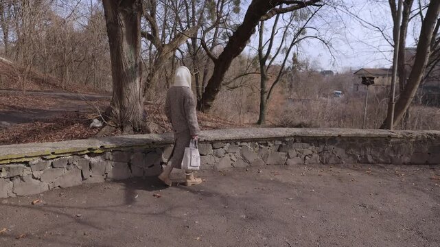 Senior woman in elegant coat and woolen scarf walks to rest on stone parapet in spring park. Healthy retired lady spends day in sunny urban garden. Activities at retirement time