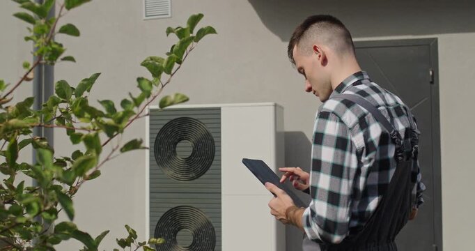 Technician checking heat pump system outdoors using tablet near residential house