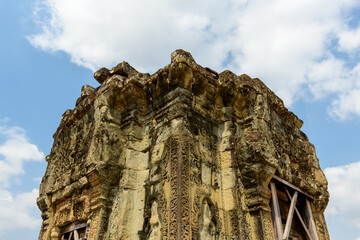 Fototapeta premium A weathered sandstone tower adorned with intricate carvings rises against a backdrop of blue sky and scattered clouds at Phnom Bakheng temple near Siem Reap, Cambodia. The historic structure displays