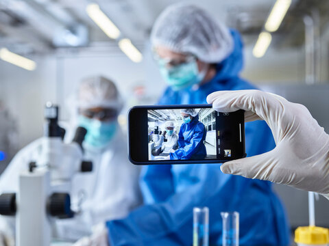 Hand holding smartphone, photographing chemists, working in industrial laboratory