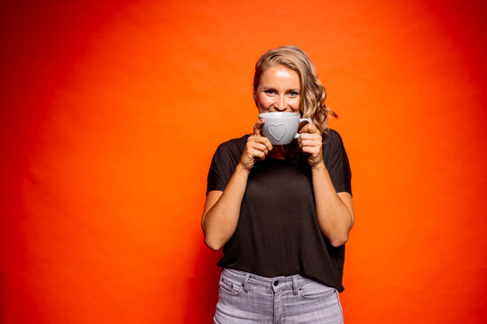 Happy woman holding coffee cup with heart shape while standing against orange background