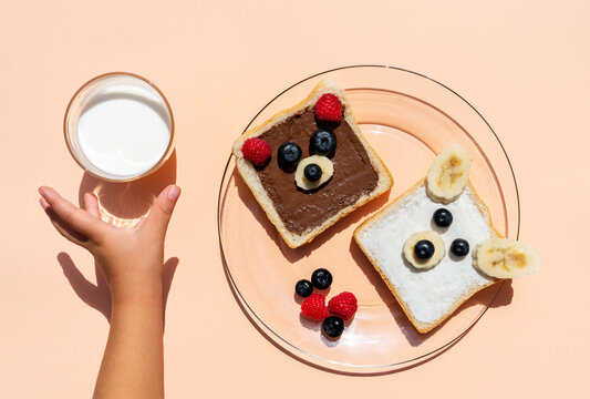 Studio shot of toasts with bear faces made of fruits and hand of baby girl reaching for glass of milk