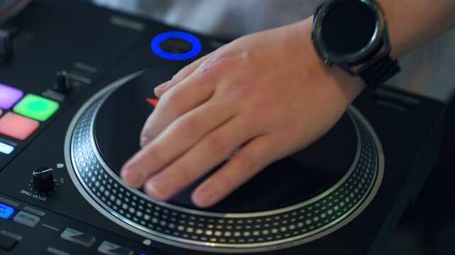 Extreme close-up of professional DJ hand scratching vinyl on black digital turntable platter with glowing blue jog wheel, colorful performance pads and waveform display during live set