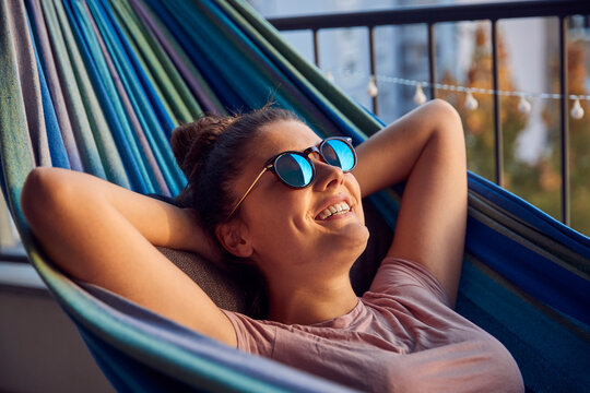 Portrait of happy young woman with sunglasses lying on hammock on balcony