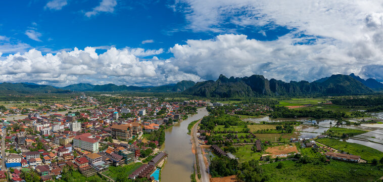 Wide panoramic view of Vang Vieng, Laos, with the Nam Song River winding through the town and dramatic limestone cliffs rising in the background. The scene features clustered buildings, green rice