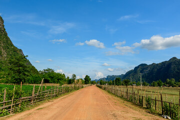 Fototapeta premium A straight red earth road runs through open farmland bordered by rustic wooden fences, with green fields and distant limestone hills beneath a vivid blue sky. The landscape features natural textures