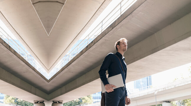 Businessman standing at underpass holding laptop, composite