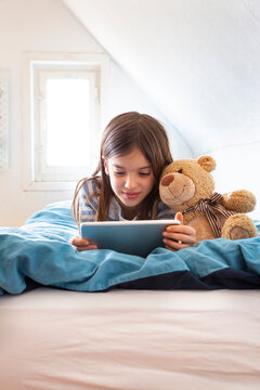 Portrait of smiling girl lying on bed with teddy bear using digital tablet