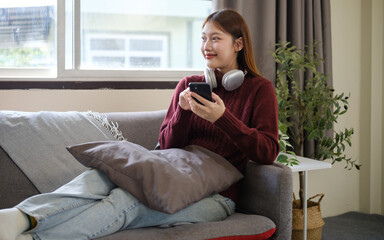 Obraz premium Young woman relaxing on a sofa while using her smartphone with headphones around her neck, enjoying a peaceful moment at home.