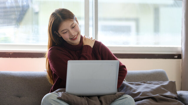 Young woman rubbing her shoulder while working on a laptop on the sofa at home. Concept of remote work, comfort, and muscle relief.