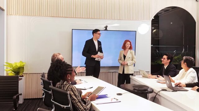 Team meeting in a modern conference room as two colleagues present by a large screen while others take notes on laptops and tablets.