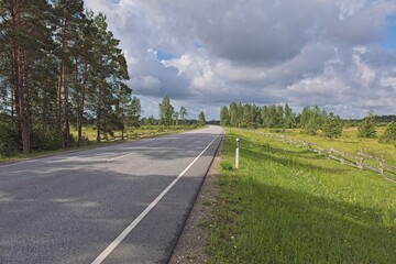 Fototapeta premium Scenic rural Kuivastu to Kuressaare road, featuring paved two lane asphalt road with traditional wooden fences lining the grassy fields alongside it in cloudy summer weather, Saaremaa, Estonia, Europe