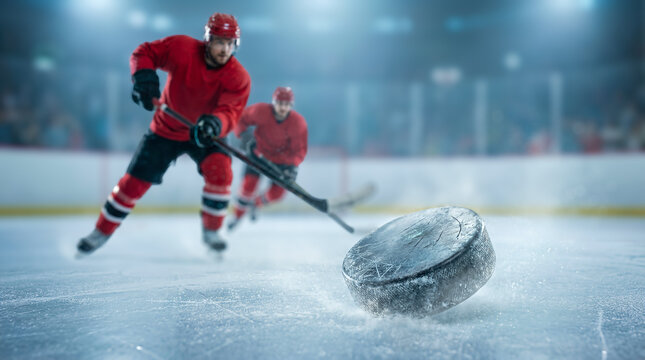Hockey players in red jerseys skating on ice rink, one player striking a puck with a stick while another player follows closely behind in a competitive setting