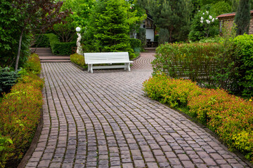 A paved curved alley and a bench in a landscape park among trimmed green bushes. Resting place, public park in the city.