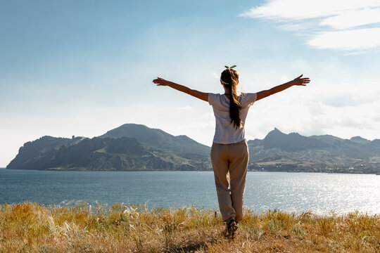 Happy young woman is standing with open arms against sea bay and sunset sky